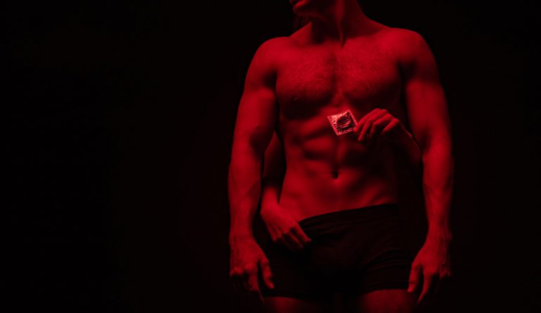 A woman's hands are wrapped around a man who has no shirt on, she is holding a condom wrapper