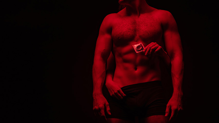 A woman's hands are wrapped around a man who has no shirt on, she is holding a condom wrapper